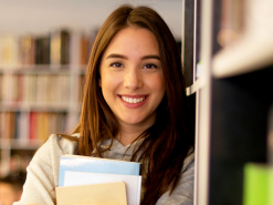Lächelnde junge Frau steht in einer Bibliothek zwischen Regalen und hält mehrere Ordner und Unter